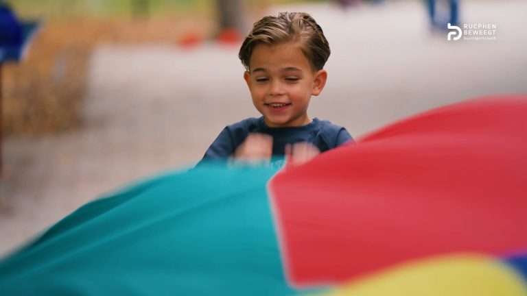 Lachende jongen speelt met een kleurrijk parachutedoek tijdens een beweegactiviteit van de buurtsportcoach