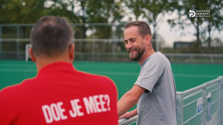 Buurtsportcoach met de tekst Doe je mee? op zijn jas in gesprek met een partner bij een sportveld